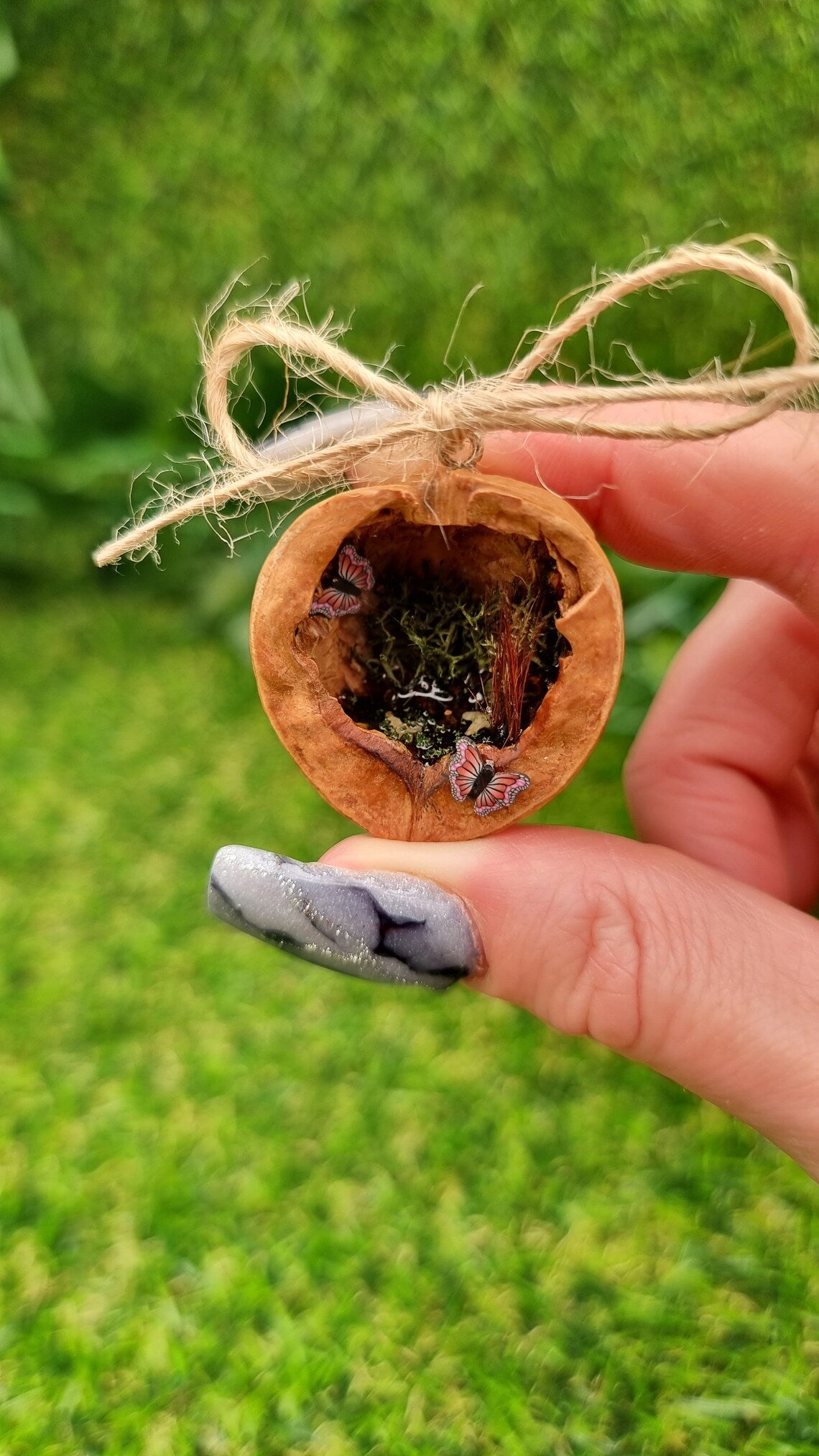 A hand holds a small brown walnut half shell containing a miniature diorama. The inside of the shell is natural brown with greenery, brown reeds and a tiny pond with leaves floating on the surface. There are two pink and purple butteflies