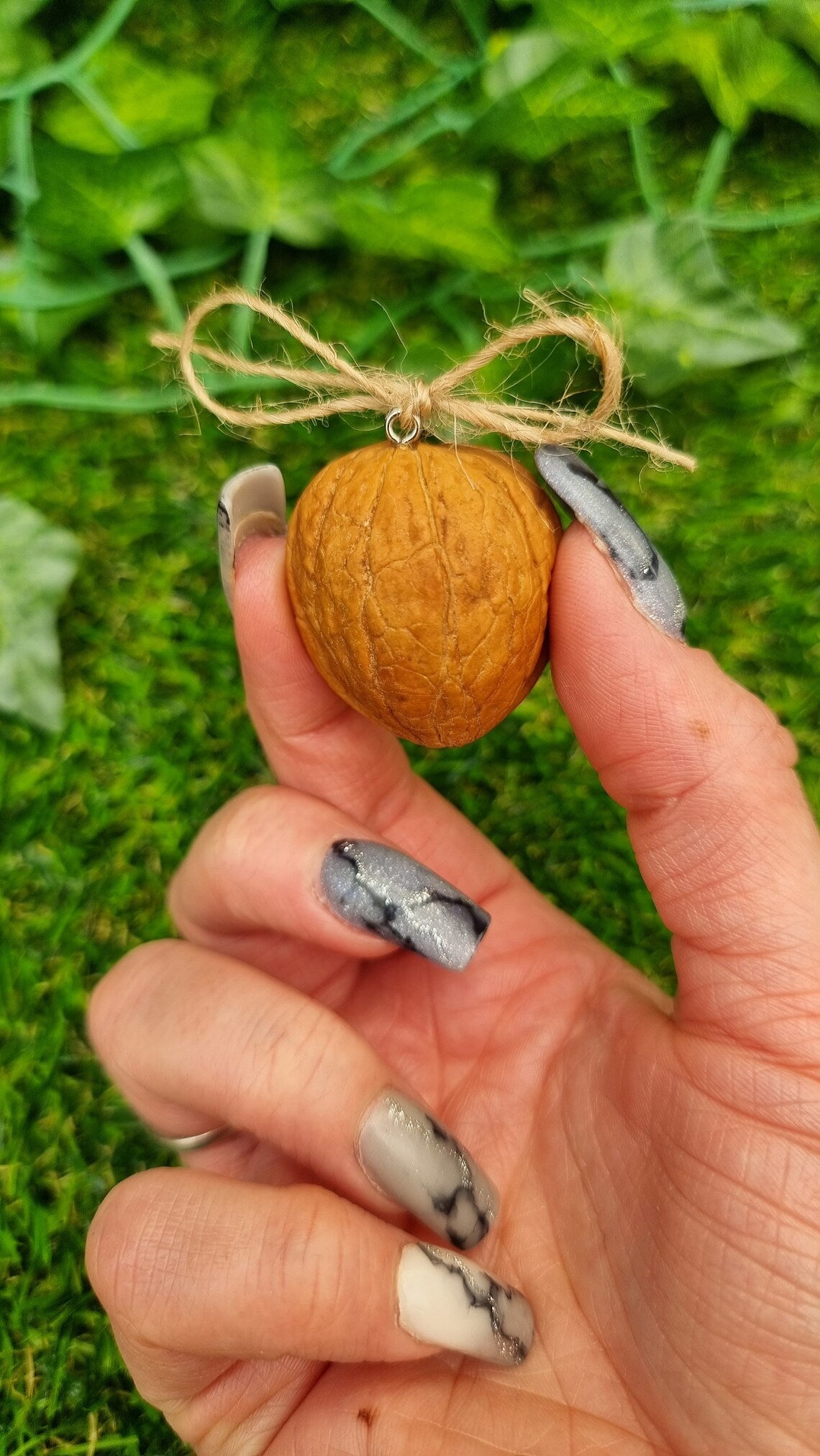 A hand holds a small brown walnut half shell containing a miniature diorama. The back of the nut is shown, with a jute string bow tied to a hook eye in the top