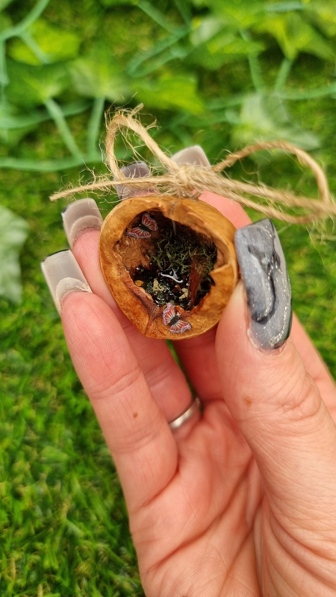 A hand holds a small brown walnut half shell containing a miniature diorama. The inside of the shell is natural brown with greenery, brown reeds and a tiny pond with leaves floating on the surface. There are two pink and purple butteflies