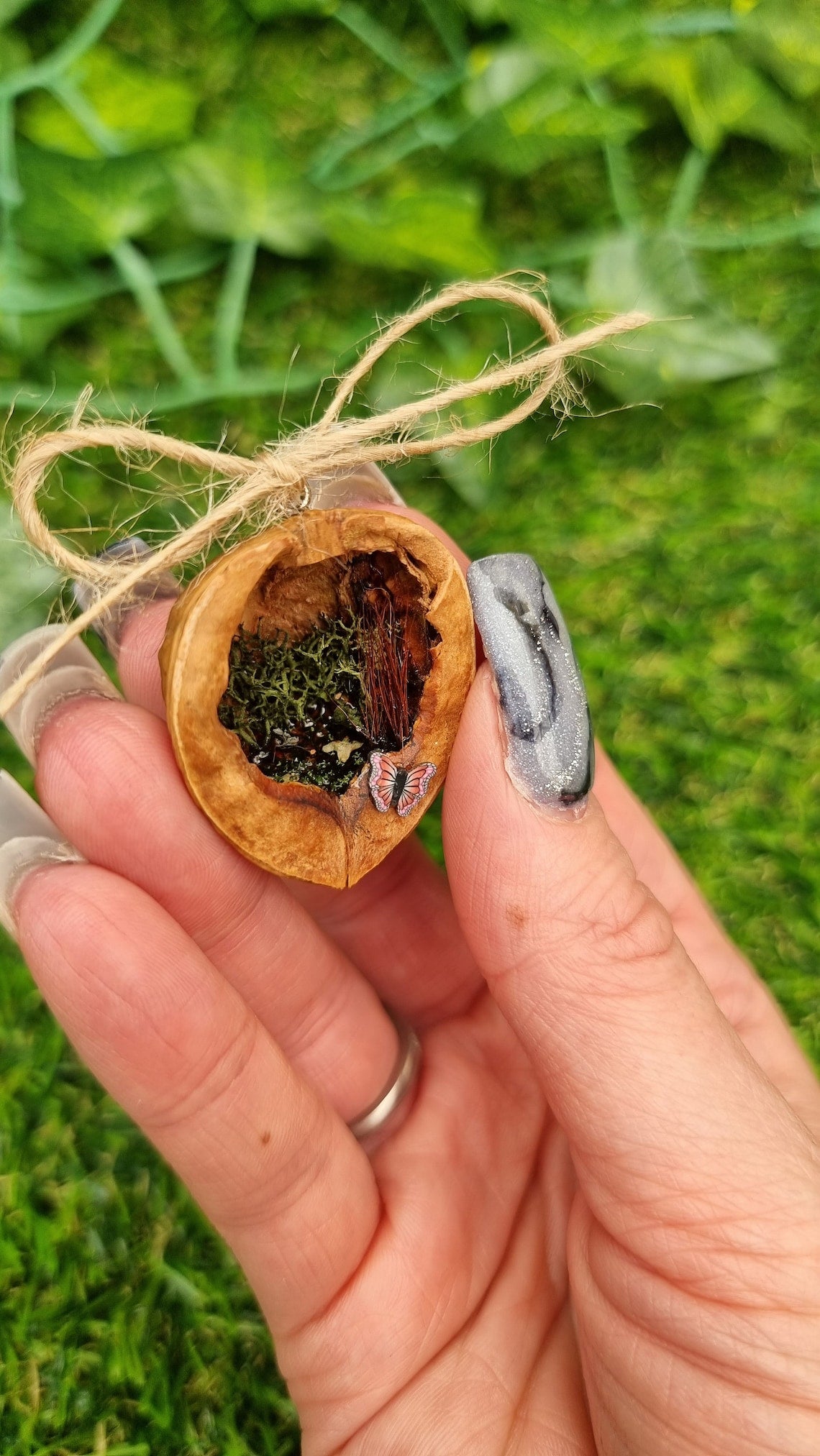 A hand holds a small brown walnut half shell containing a miniature diorama. The inside of the shell is natural brown with greenery, brown reeds and a tiny pond with leaves floating on the surface. There are two pink and purple butteflies