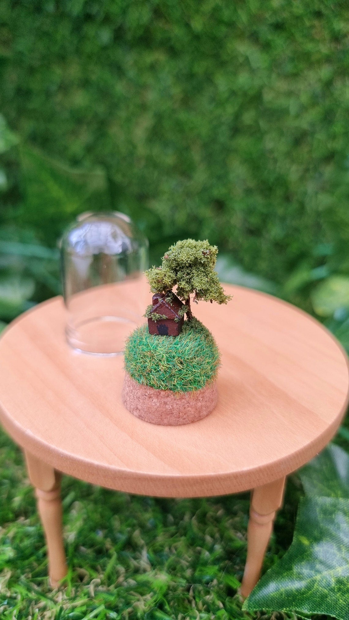 Miniature cork base containing a tiny brown house and a tree on top a grassy hill sits on a 1:12 scale table. There is a small glass bell jar lid sat behind it