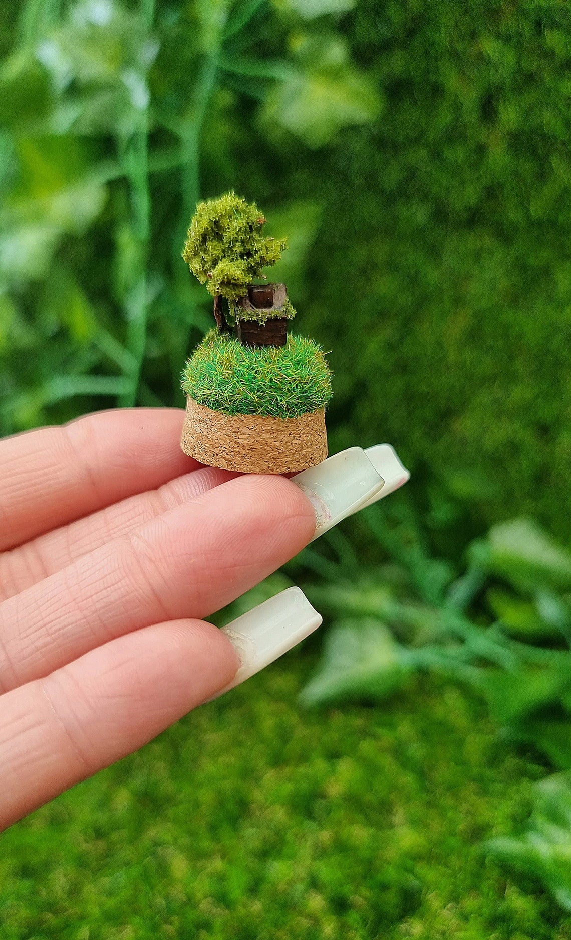 Side view of a miniature diorama on a cork base. It has a tiny brown house and a tree on top a grassy hill sits on a 1:12 scale table. There is a small glass bell jar lid sat behind it