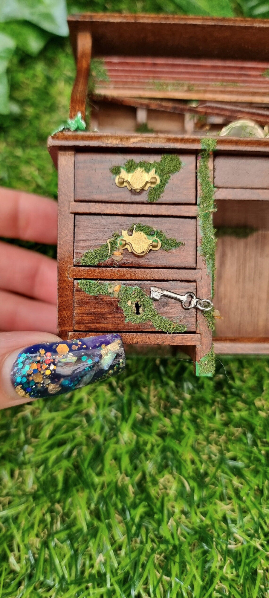 Front of a 1:12 scale miniature abandoned rolltop desk. The desk is brown with green moss and algae creeping across 3 drawers. one of the drawers does not have a handle and has a keyhole with a key beside it