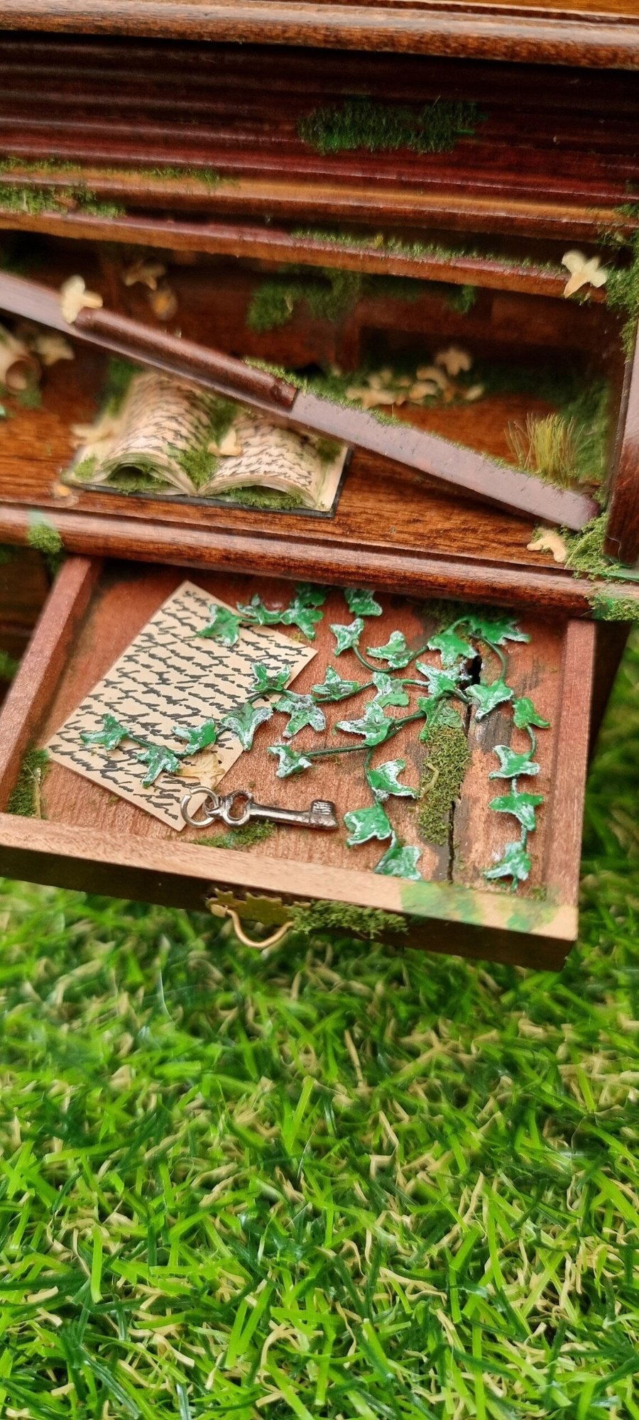 1:12 scale miniature abandoned rolltop desk with the top drawer open.  The drawer contains green creeping ivy, moss, an old letter and a silver key. The desk top has a mossy old book, leaves and a broken wooden slat of the roll top