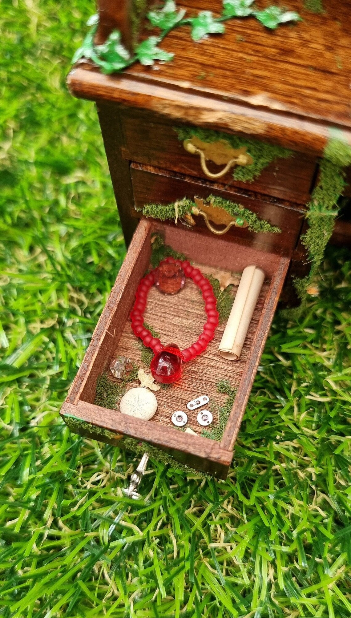 1:12 scale miniature abandoned desk drawer is open, revealing moss, a sea urchin, dead leaves, trinkets and a red bead necklace. A silver key sticks out from the front of the drawer