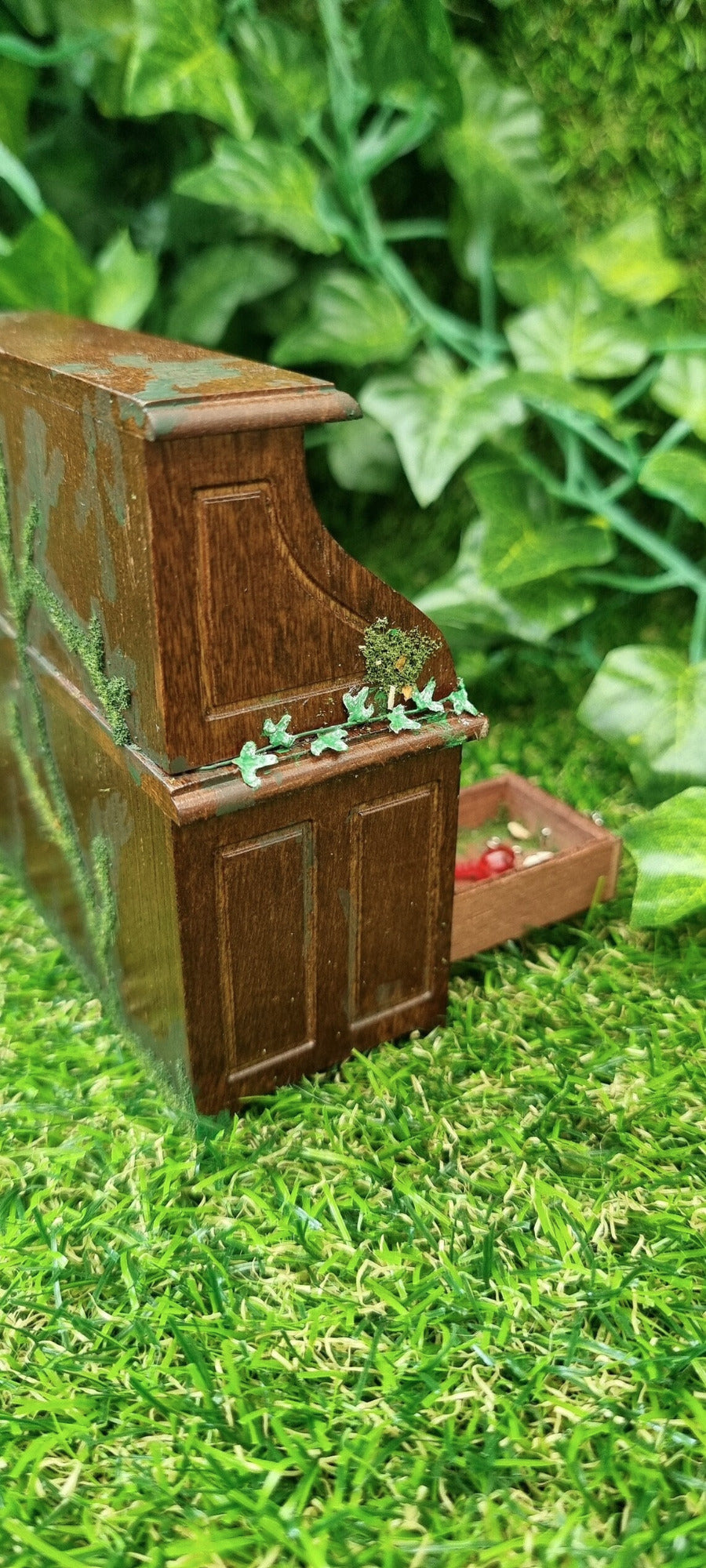 Side of a miniature 1:12 scale abandoned rolltop desk. The desk is brown with green moss and algae creeping across the back. Green ivy is creeping from the back to the front of the desk. It sits against a green grass background.