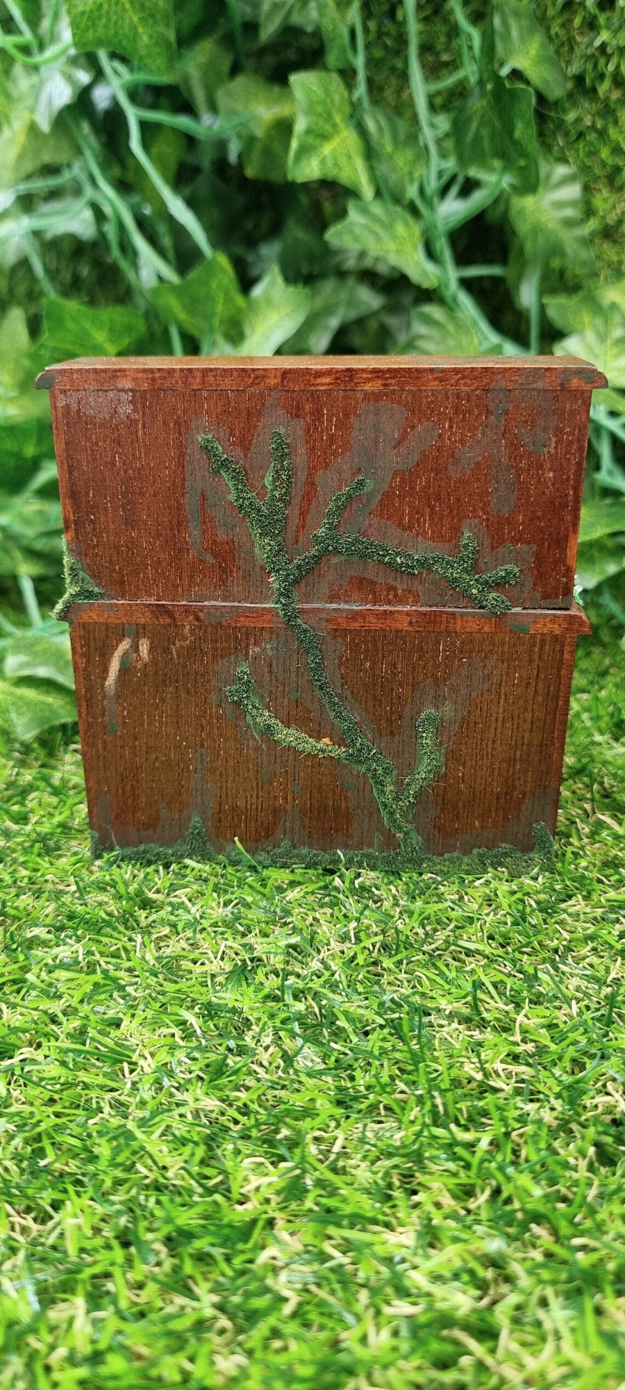 Back of a 1:12 scale miniature abandoned rolltop desk. The desk is brown with green moss and algae creeping across it. The desk is shown against a green grass background