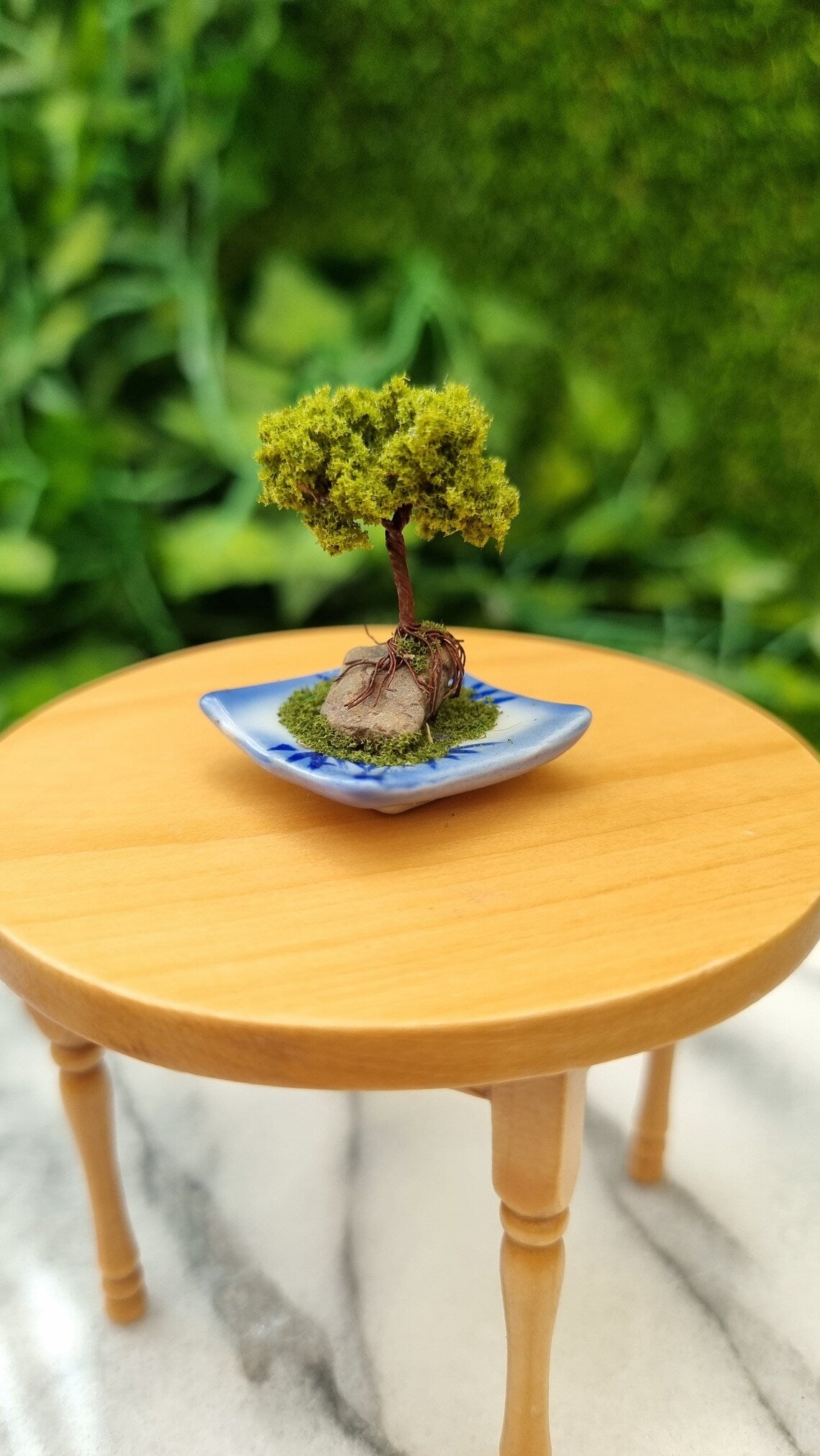 1:12 scale bonsai tree sits on a miniature table. The bonsai is mounted on a small stone on a blue and white square plate with moss