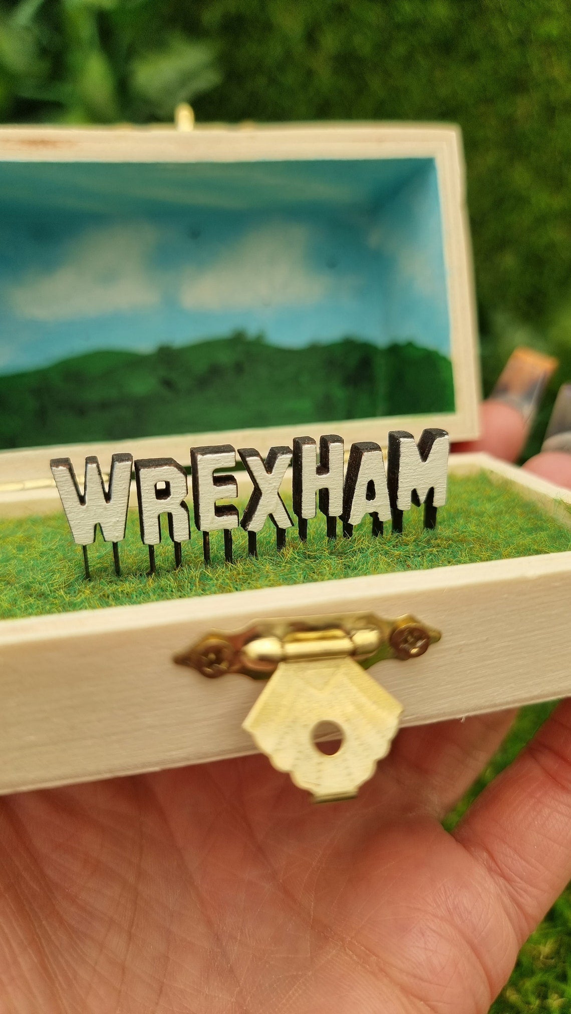 A small hinged wooden box sits open. The box contains a white hollywood-style Wrexham sign on flocked grass field. The deep lid of the box sits behind with a painted countryside scene with green hills, blue sky, white clouds and soaring birds