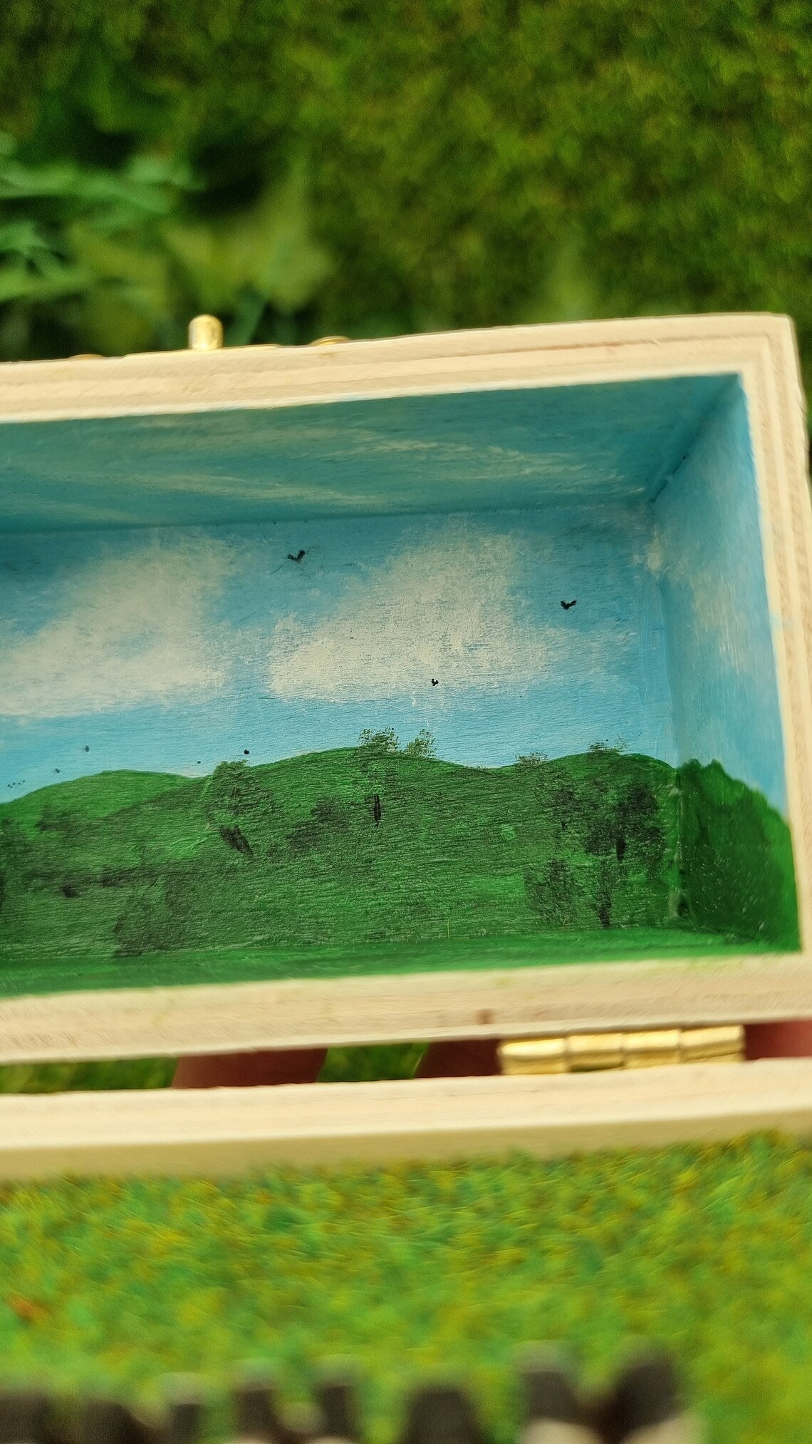 Details of a small open hinged wooden box diorama. The deep lid of the box has a painted countryside scene with green hills, blue sky, white clouds and soaring birds