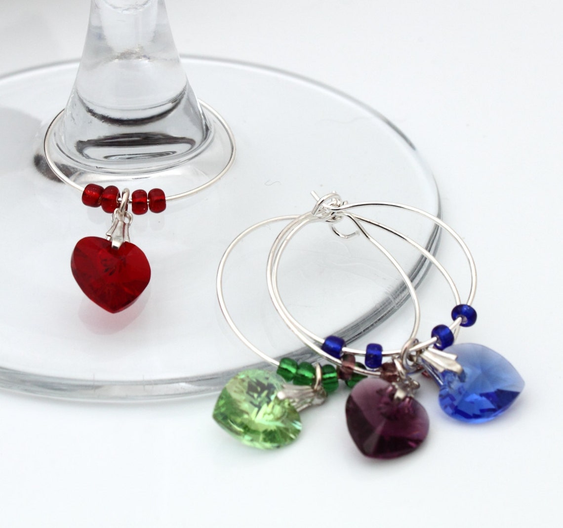 3 wine glass charms are arranged on top of  wine glass bases on a wooden background. The wine glass charms are round wire hoops, each with different coloured seed beads and a Swarovski crystal heart in red, green and lavender