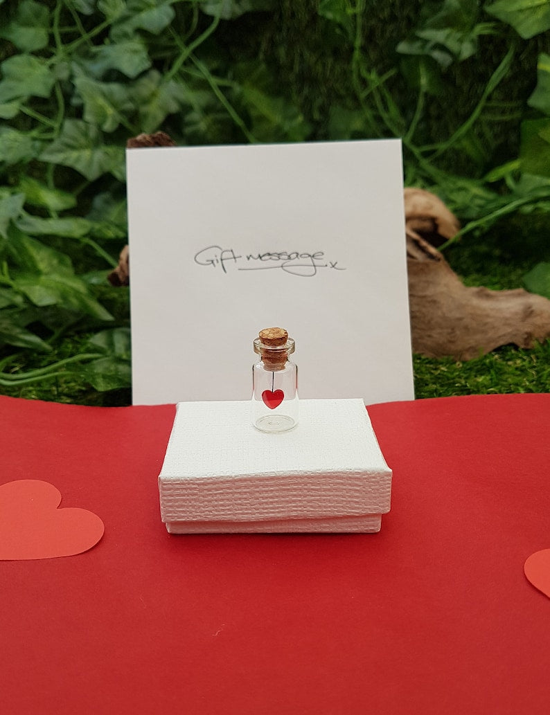 A miniature glass jar with cork lid is sat on a white gift box on a red background. The jar has a red sequin heart hanging from the cork inside the jar. A gift message envelope is seen behind