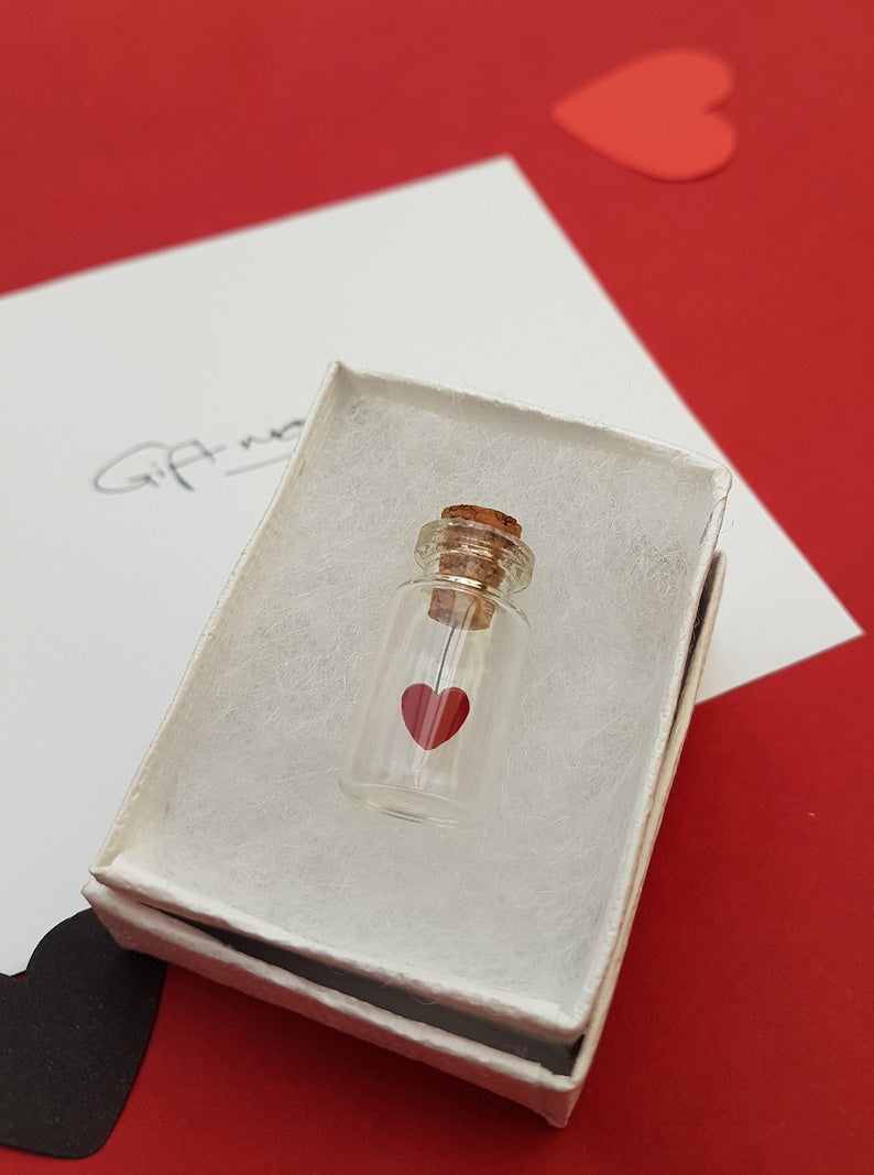 Small glass jar with cork lid with a red sequin heart hanging from the cork inside the jar. The jar is inside a white gift box