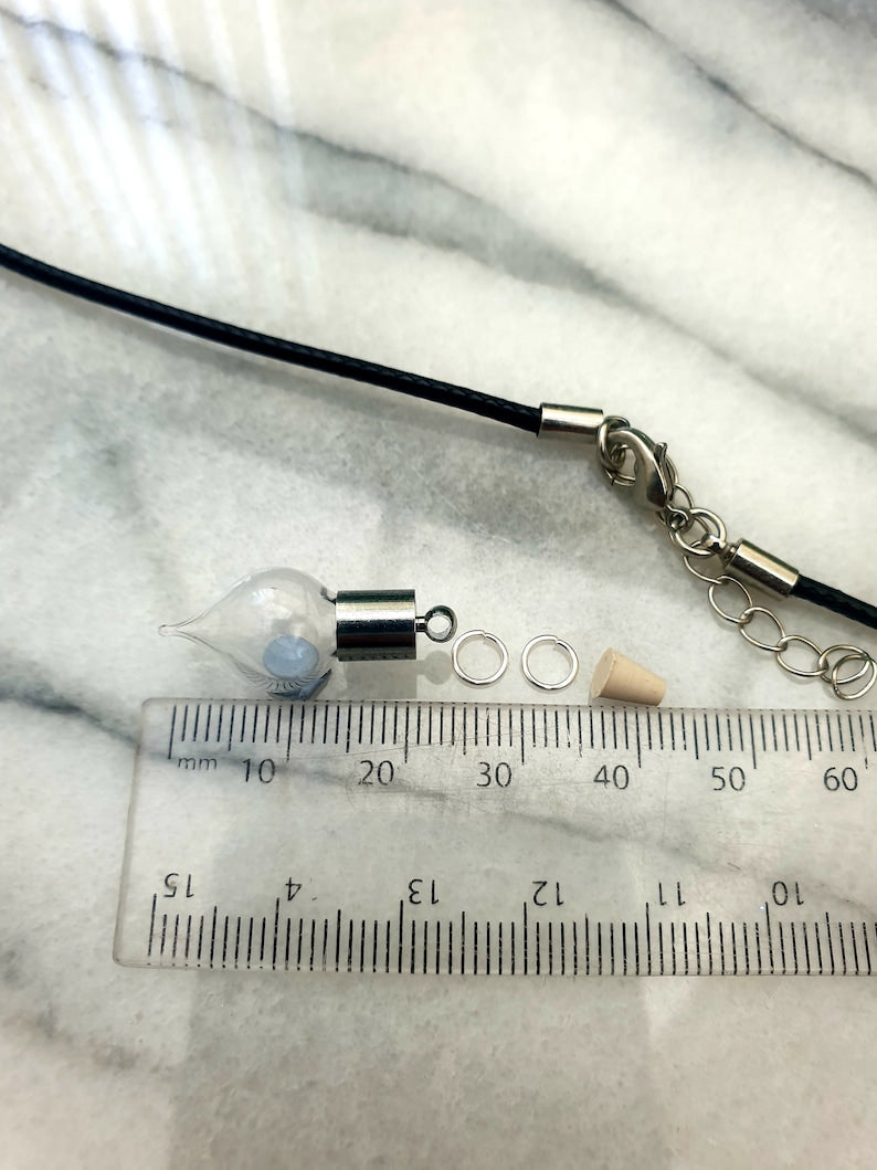 A miniature glass vial with silver coloured caps shown on a white marble slab with a ruler for scale. The vial looks like an amphora, rounded with a pointed base. 2 jump rings, a rubber bung and a black pendant cord seen in the background.