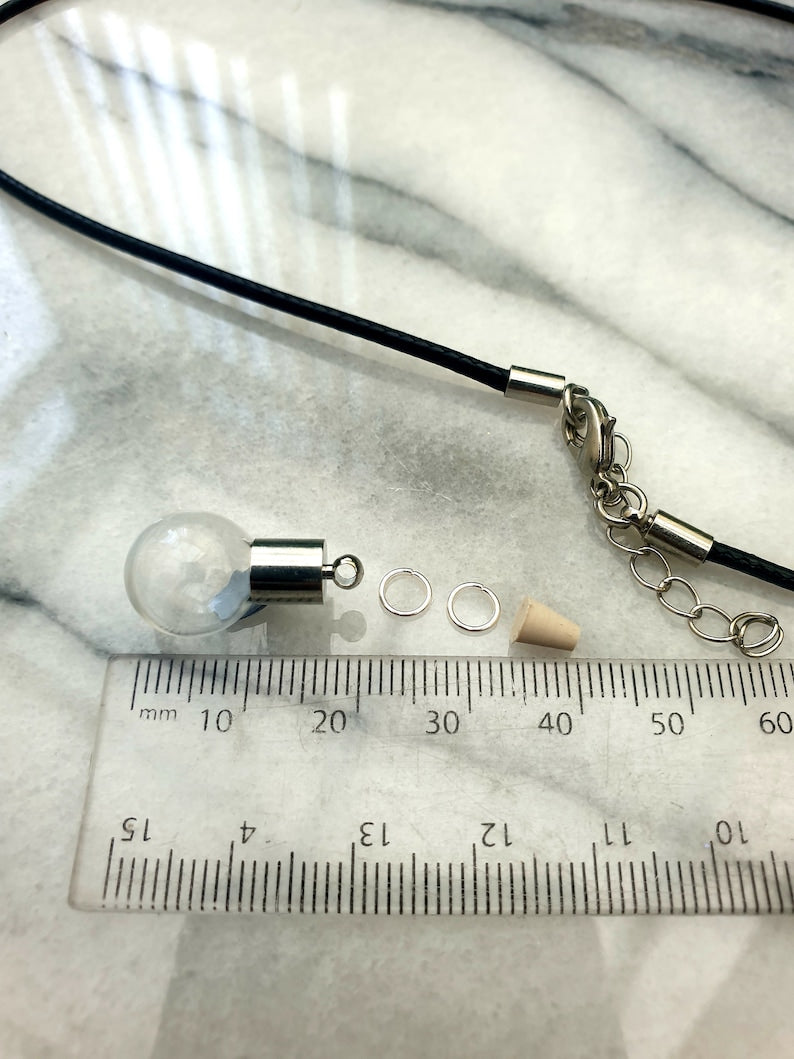 A miniature glass globe shaped vial with silver coloured cap is shown on a white marble slab with a ruler for scale. 2 jump rings, a rubber bung and a black pendant cord seen in the background.