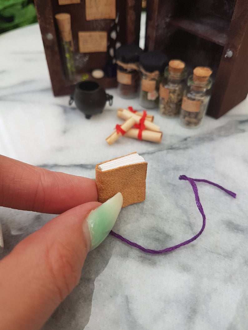 A 1:12 scale miniature brown leather bound book is held between fingers and thumb with an apothecary cabinet and contents seen in the background, including glass vial, cauldron and brown parchment scrolls