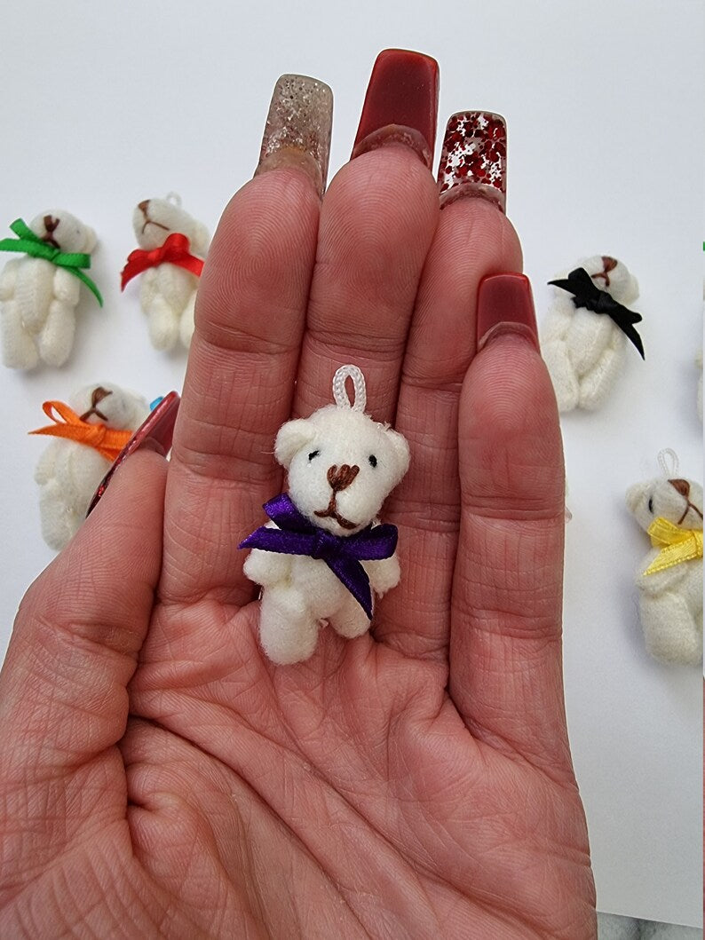 Hand holds a single miniature cream Teddy bead with a purple ribbon bow tie. Other cream bears with different colour bow ties can be seen in the background on white