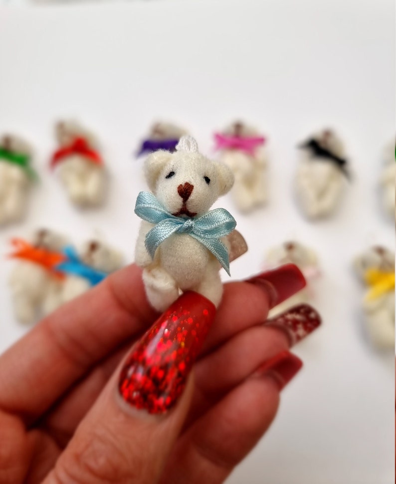 Single miniature cream Teddy bead with a pastel blue ribbon bow tie held between finger and thumb. Other cream bears with different colour bow ties can be seen in the background on white