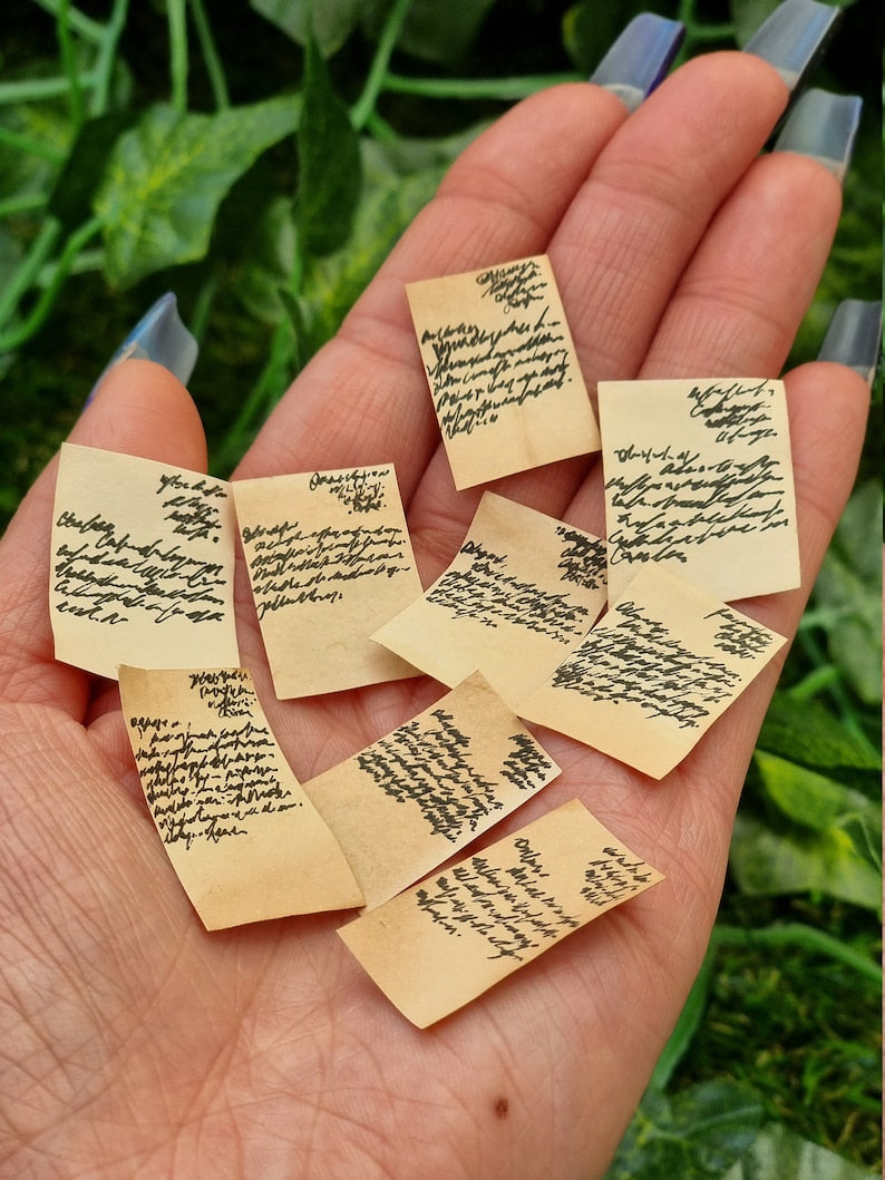 Lots of aged 1:12 scale miniature letters are displayed in the open palm of a hand. The letters are brown, showing black text. The letters are half written. Green ivy is seen in the background