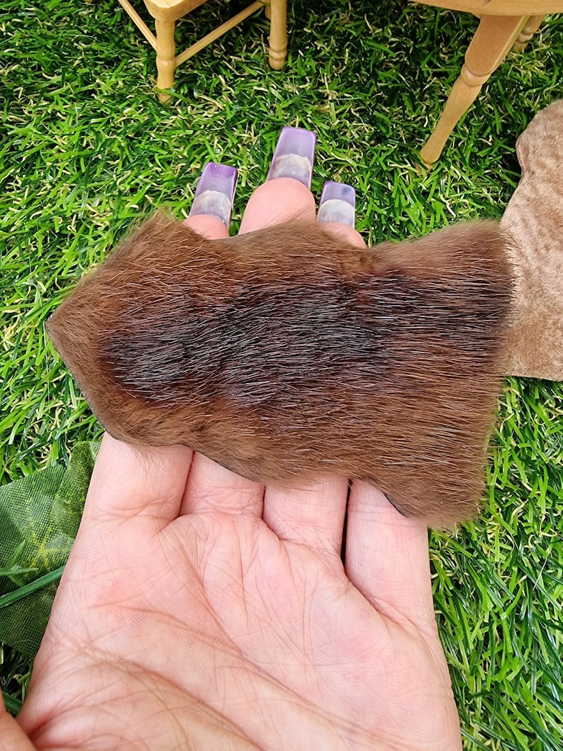 Hand holds a 1:12 scale dark brown animal skin genuine fur rug on a green grass floor in the background. The rug is dark brown with lighter areas to the edges