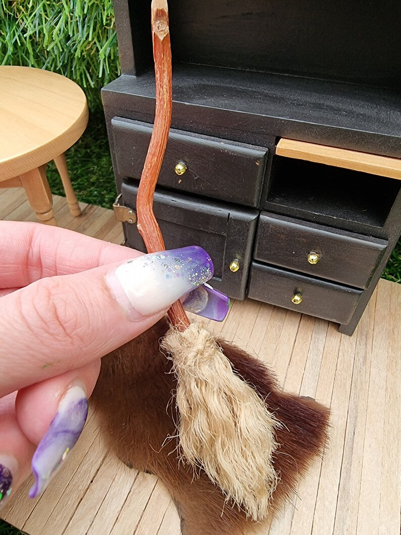 Hand holds a 1:12 broom with a dresser and rug in the background. The broom has brown bristles and a brown bent handle for flying