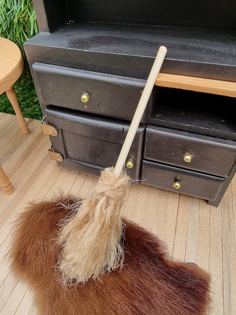 A small 1:12 broom leans against a black dresser and rug in the background. The broom has brown bristles and a long straight handle for sweeping