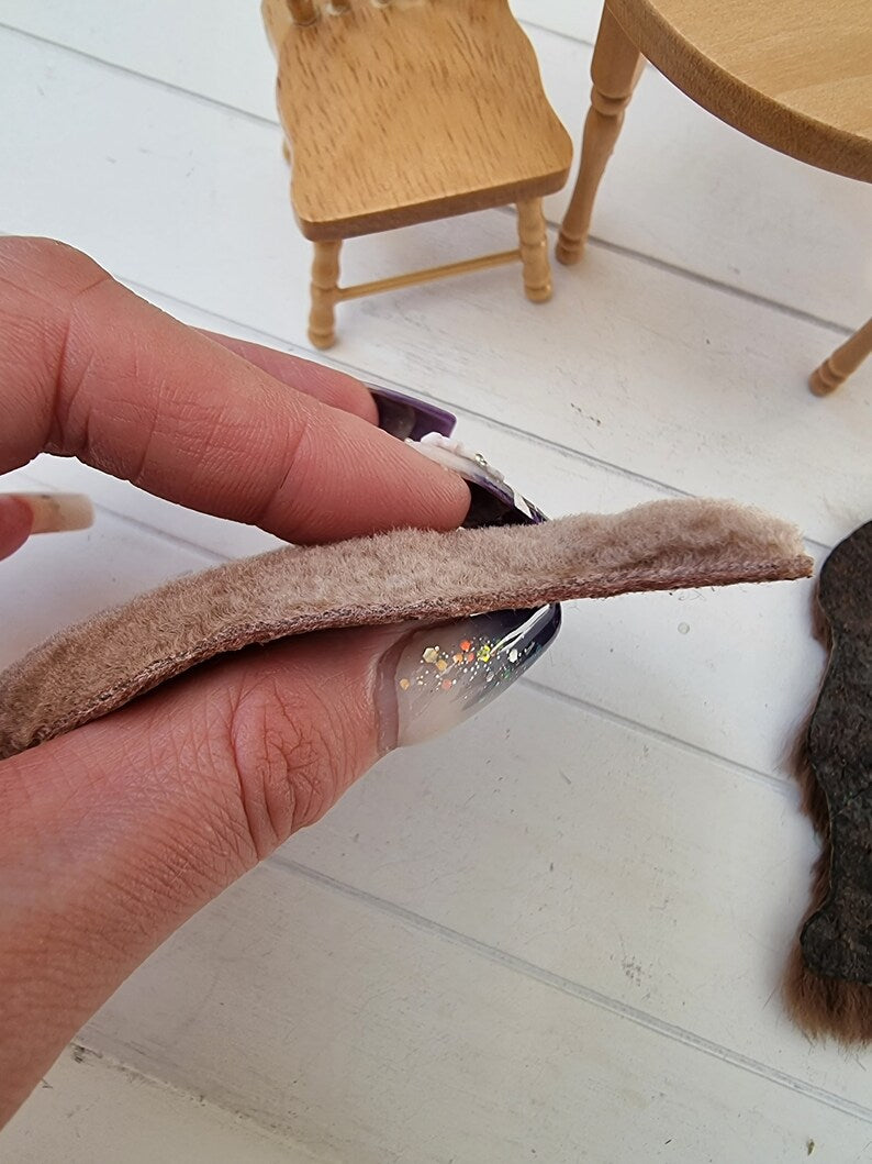 Hand holds a light brown 1:12 scale animal skin genuine fur rug to show the depth of the rug pile. The rug appears thick and deep and doesnot flatten down. Miniature furniture sits in the badkground