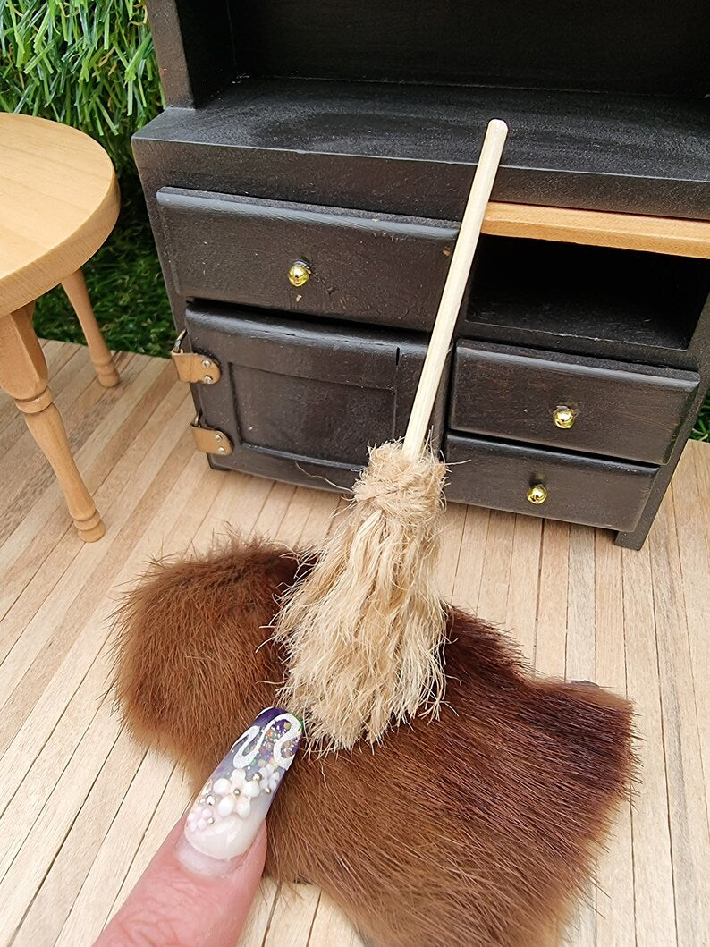 A finger points at a small 1:12 broom leaning against a black dresser and rug in the background. The broom has brown bristles and a short straight handle for sweeping