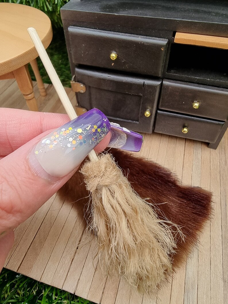 A finger points at a small 1:12 broom leaning against a black dresser and rug in the background. The broom has brown bristles and a long straight handle for sweeping