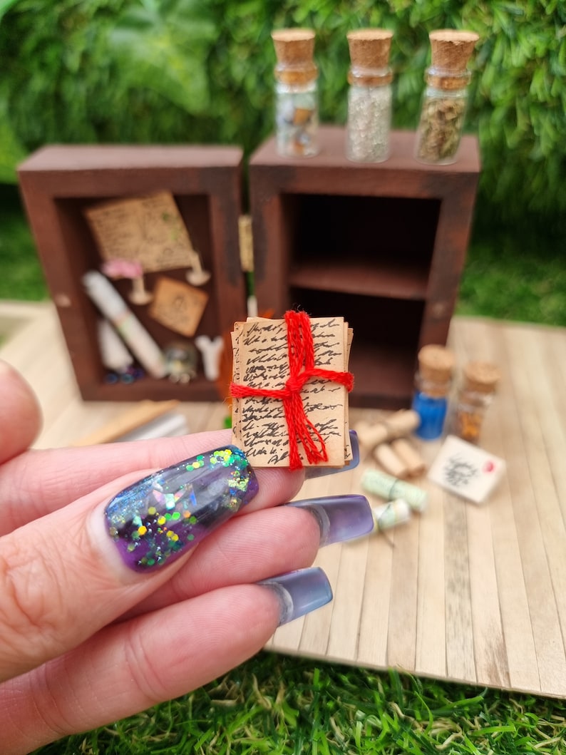 A 1:12 scale miniature brown apothecary cabinet shown open on a wood floor containing glass vials, parchment scrolls, letters, notes, inkwell and quill. A hand holds a bundle of written notes on parchment