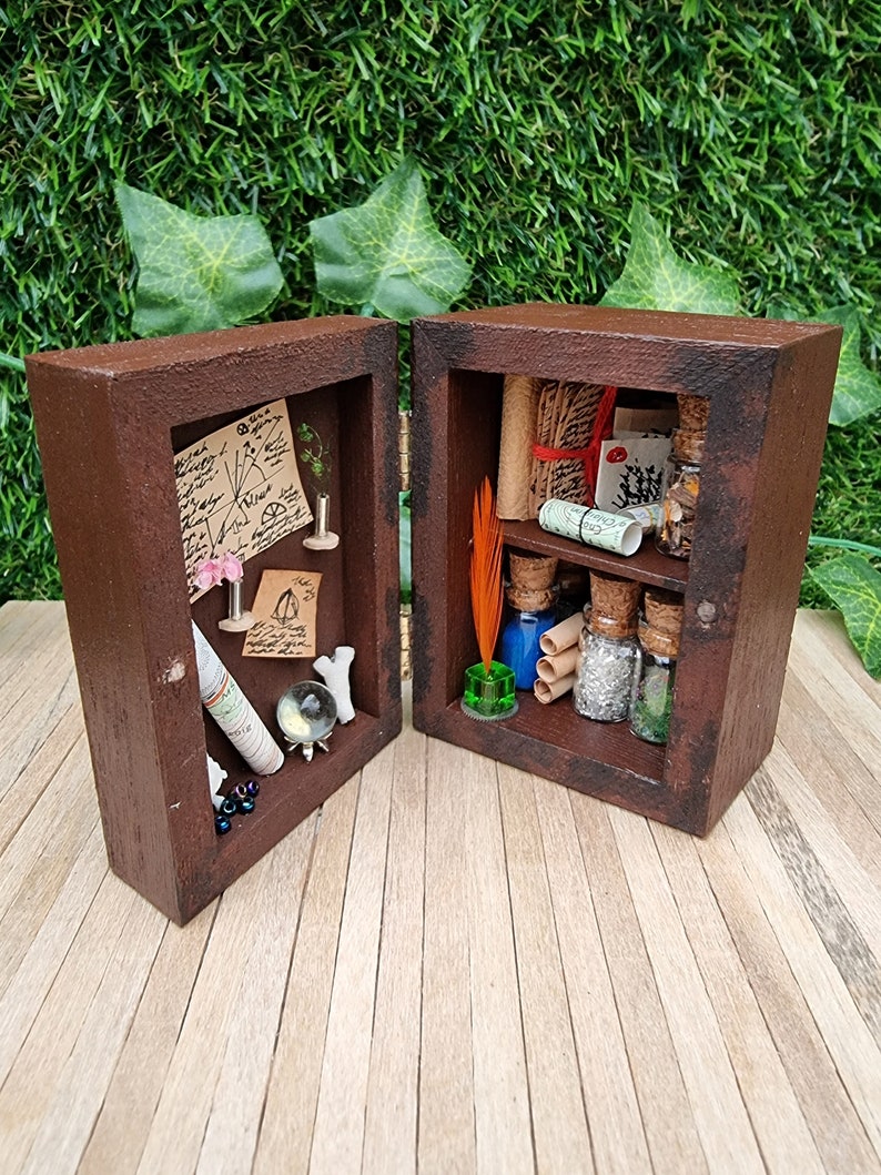 A 1:12 scale miniature brown apothecary cabinet shown open on a wood floor containing glass vials, parchment scrolls, letters, notes, inkwell and quill. The left cabinet door has aged notes pinned up, shells, crystal ball, map, coral and slim vases.