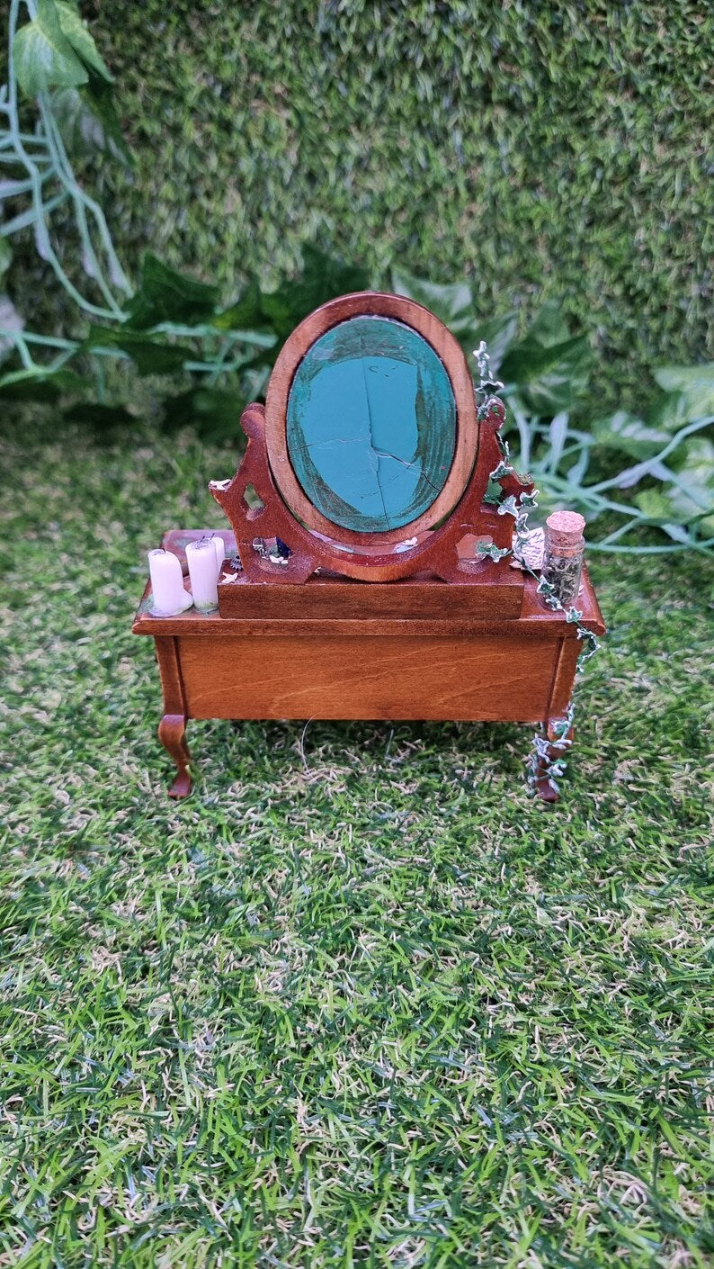 Image showing the back of an abandoned 1:12 scale dressing table. miniature ivy can be seen creeping up the back and side, a glass bottle and 2 candles can be seen either side of the damaged mirror