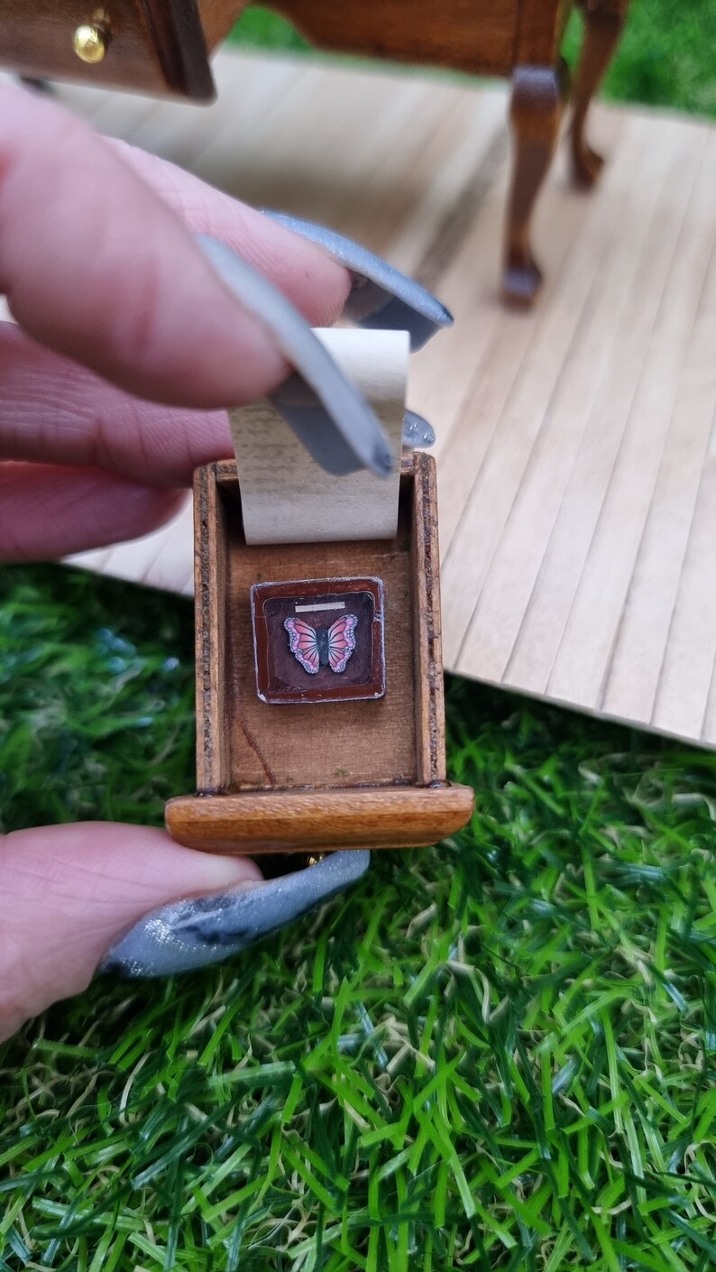 2 Fingers hold a drawer of a 1:12 abandoned dressing table. Inside is a lifted piece of paper revealing a butterfly in a box beneath. the butterfly is pink and puirple in a dark brown box with a 'glass' face and label