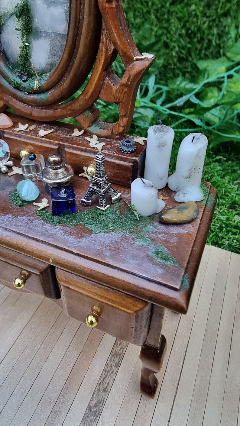 Detail of the top of a 1:12 brown abandoned dressing table. It has a a mossy broken mirror, dead leaves, strewn vessels, candles, gemstones and a minaiture eiffel tower model