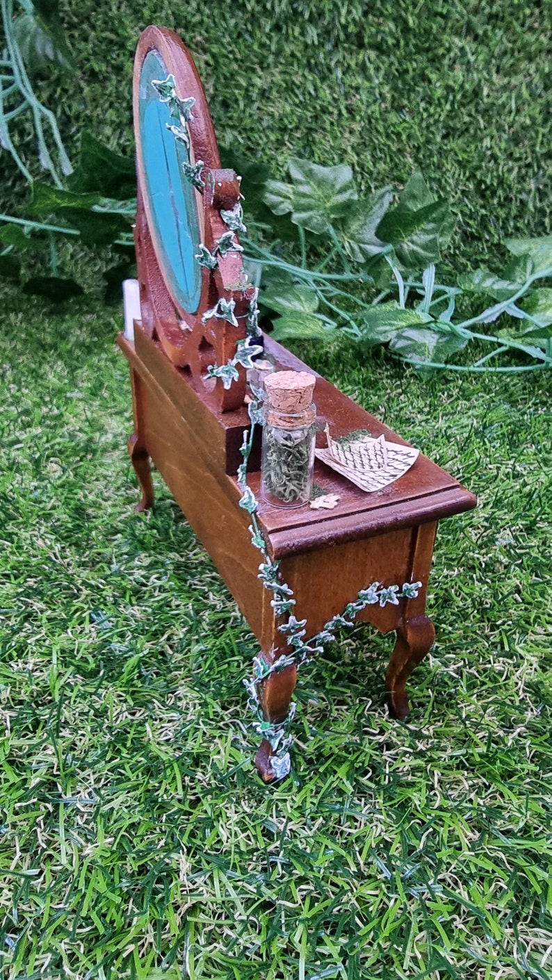 Image showing the side of an abandoned 1:12 scale dressing table. Miniature ivy can be seen creeping up the back and side, a glass bottle and handwitten notes can be seen on the dresser top and the back of the broken mirror