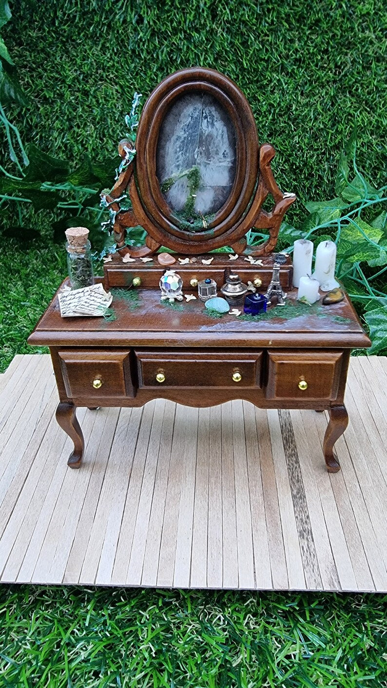 Image showing the side of an abandoned 1:12 scale dressing table. Miniature ivy can be seen creeping up the back and side, a glass bottle and handwitten notes can be seen on the dresser top and the back of the broken mirror