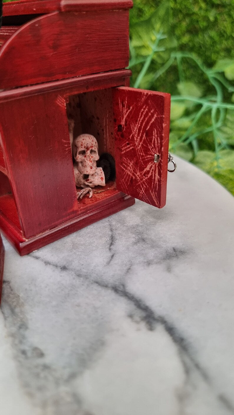 Side view of a 1:12 scale brown rolltop desk showing an open cupboard door with a keyhole in the side of the desk. There are scratches all over the inside of the door and the top left corner above the cupboard door. There are skulls and bones inside