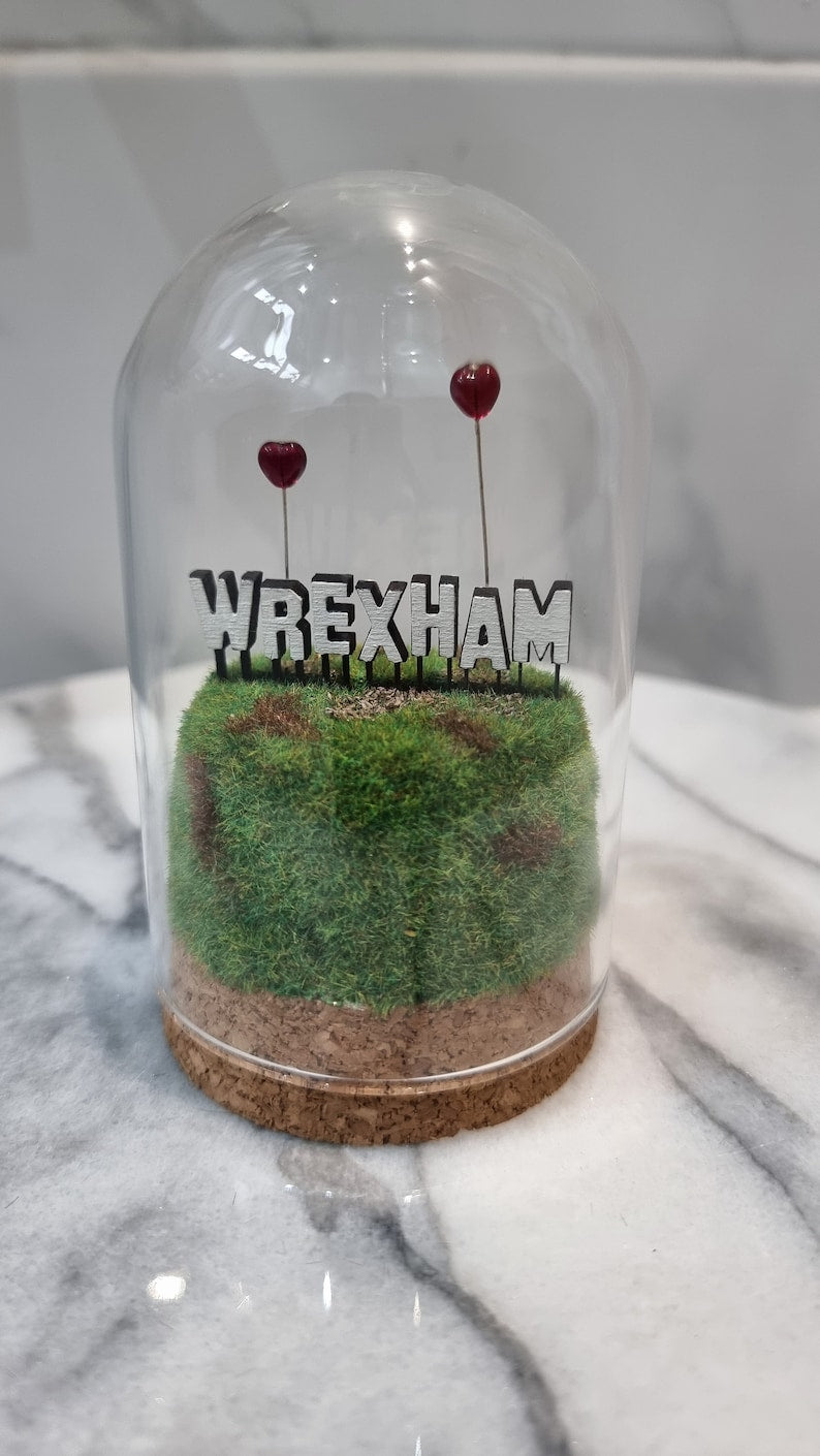 A small diorama with a glass dome cover contains a green and brown grassy hill topped with a white Hollywood-style Wrexham sign on top. The letters R and A have red heart shaped balloons on wires floating above the sign. Background of marble