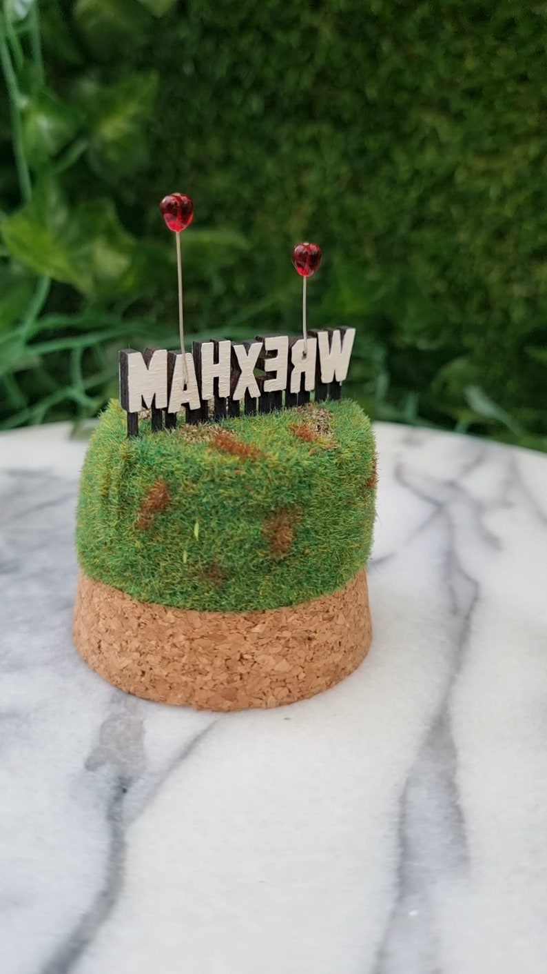 A small diorama is sat on a marble background. There is a green and brown grassy hill topped with a Hollywood-style Wrexham sign on top seen from the back. The R and A have red heart shaped balloons on wires floating above the sign