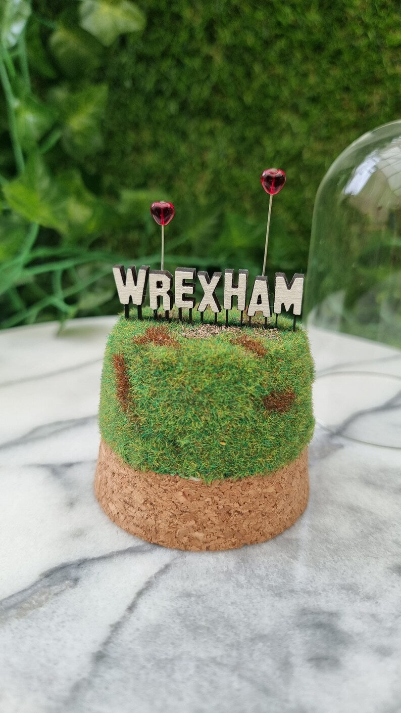 A small diorama is sat on a marble background. There is a green and brown grassy hill topped with a Hollywood-style Wrexham sign on top seen from the back. The R and A have red heart shaped balloons on wires floating above the sign