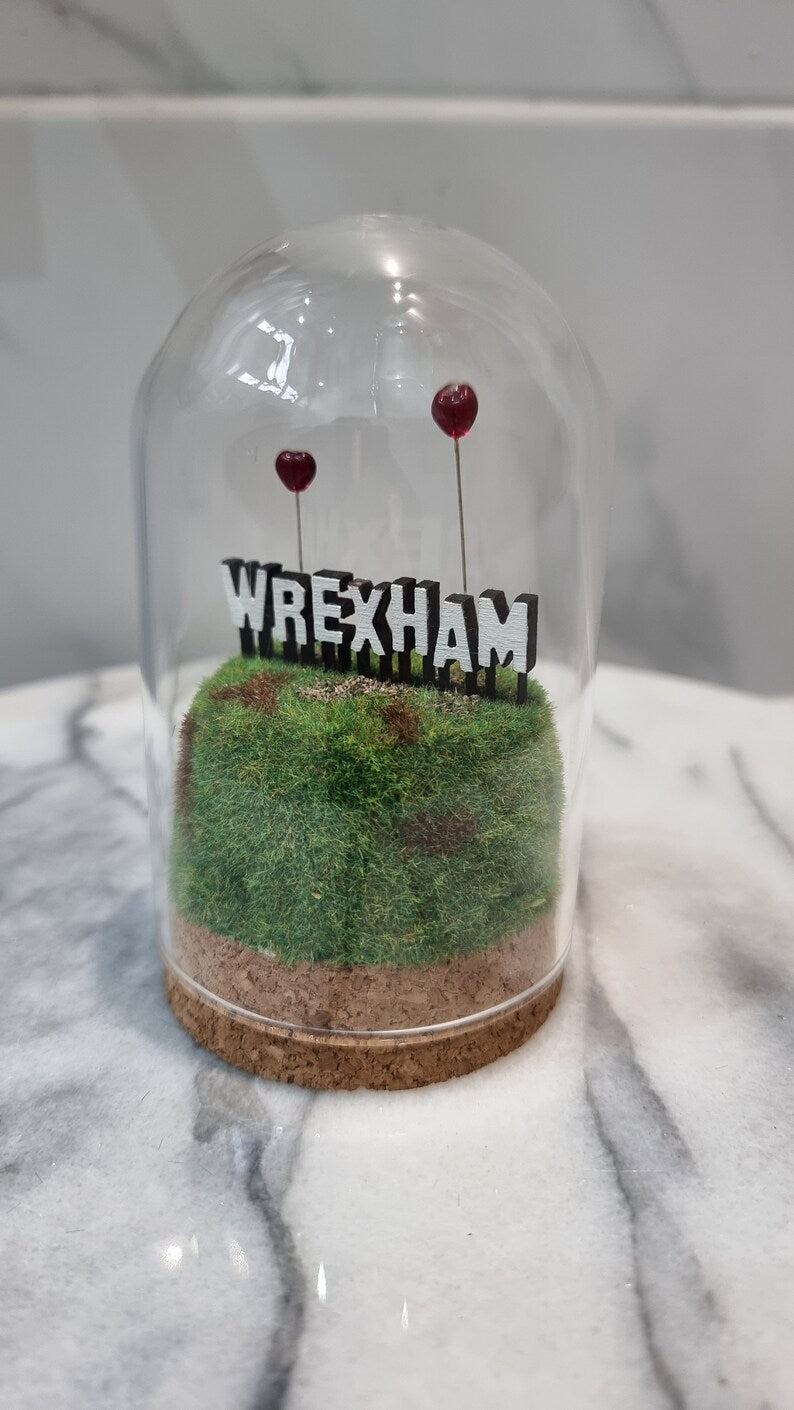 A small diorama with a glass dome cover contains a green and brown grassy hill topped with a white Hollywood-style Wrexham sign on top. The letters R and A have red heart shaped balloons on wires floating above the sign. Background of marble