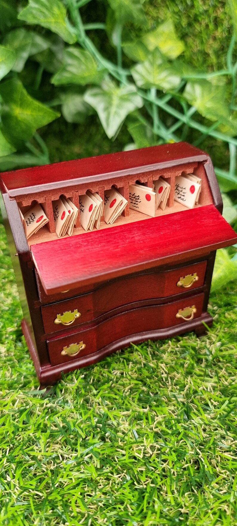 A 1:12 scale brown miniature bureau desk sits against a green background. The desk is topped with stacks of old letters, inkwell and quill. Two drawers are open, revealing papers, candles, map, curios ad trinkets