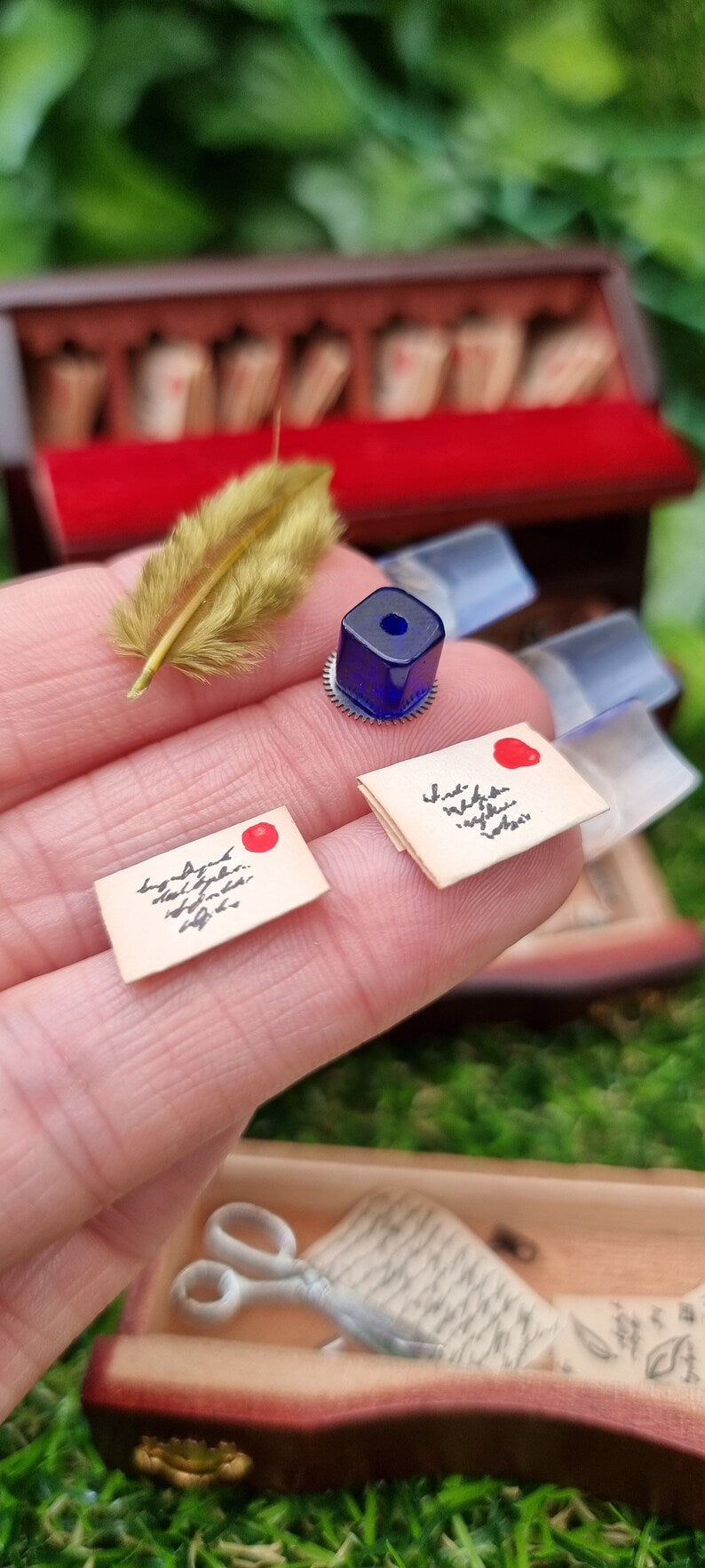 Tiny 1:12 scale feather quill, square blue inkwell and miniature letters sit on fingertips held in front of a miniature bureau desk. The miniature desk in the background has an open top with letters. A drawer containing scissors and papers is shown