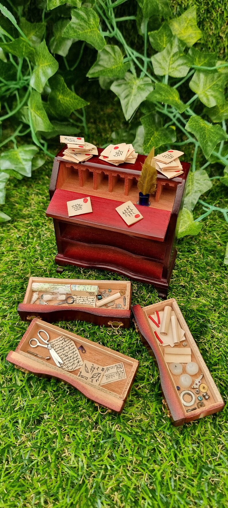 A 1:12 scale brown miniature bureau desk sits against a green background. The desk top is open, showing an inkwell and old letters stacked on top. Three drawers are out in front of the desk, revealing papers, candles, map, curios and trinkets