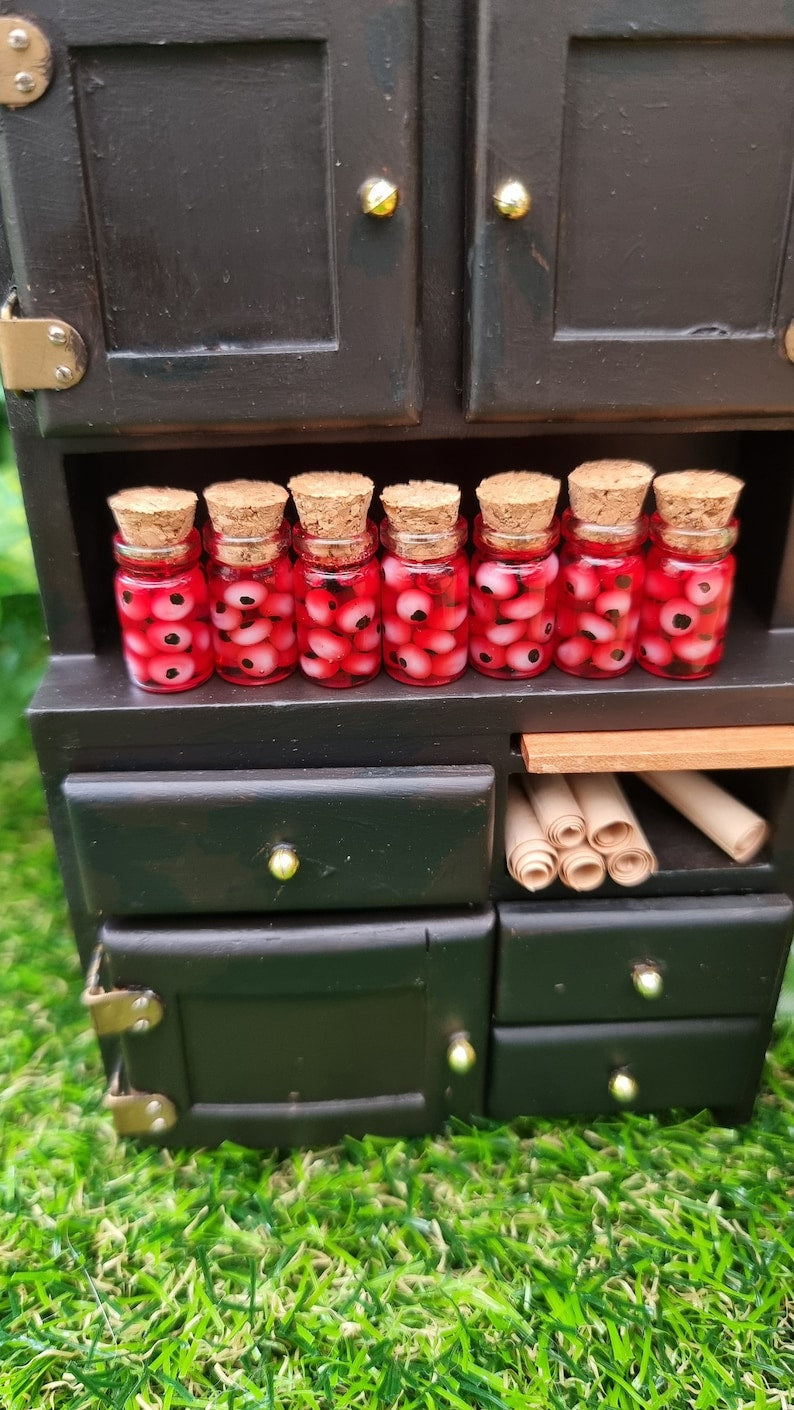 A 1:12 miniature black dresser holds 7 miniature glass jars containing eyeballs and red liquid.  The miniature jars are topped with cork stoppers