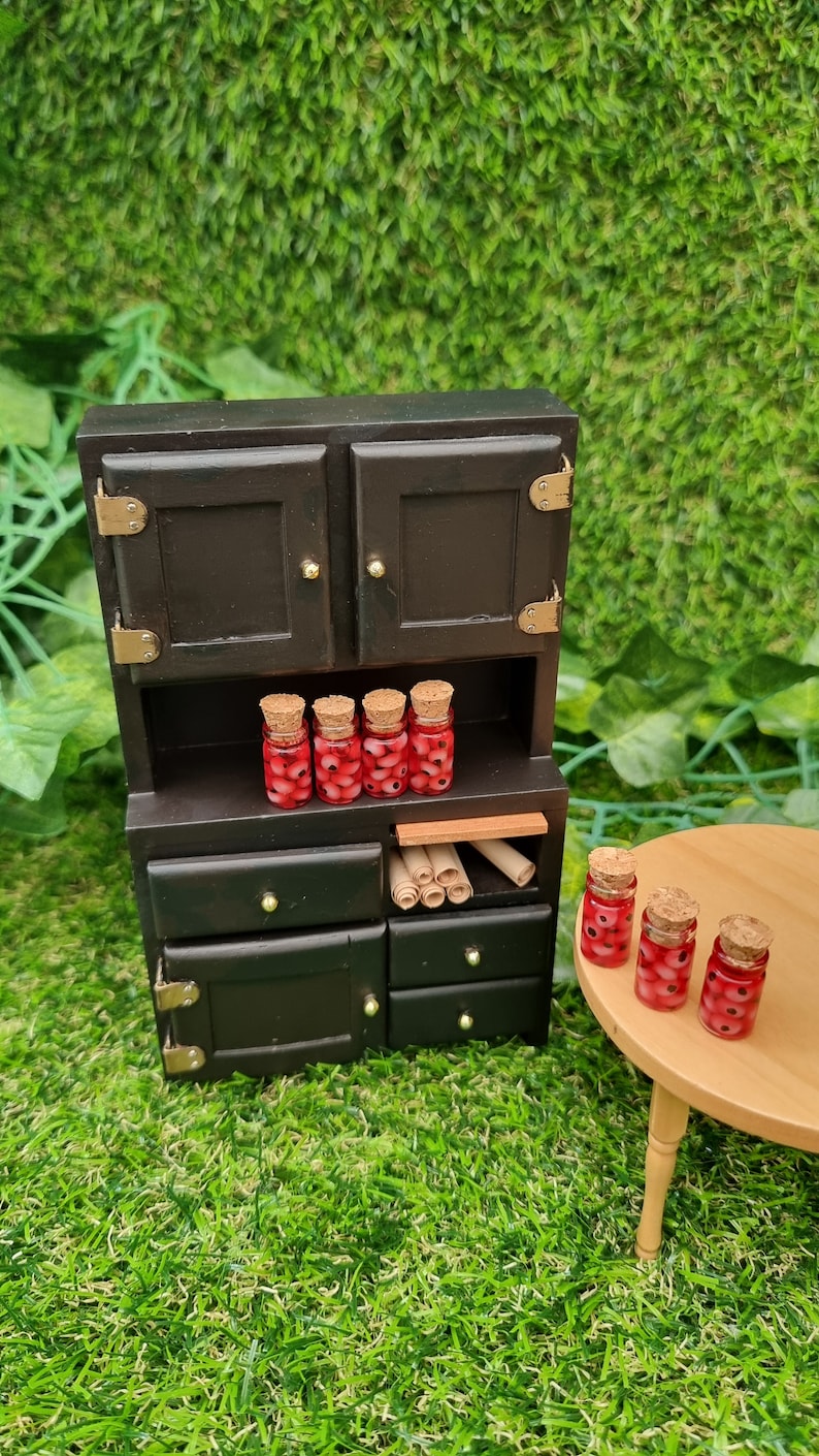 A 1:12 miniature black dresser  and round wooden table holds 7 miniature glass jars containing eyeballs and red liquid.  The miniature jars are topped with cork stoppers