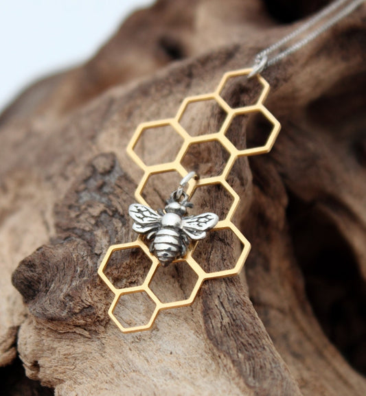 A sterling silver bee charm hangs from the centre of a brass geometric filigree honeycomb piece. The silver bee compliments the golden brassl the charms hangs on a fine sterling silver chain