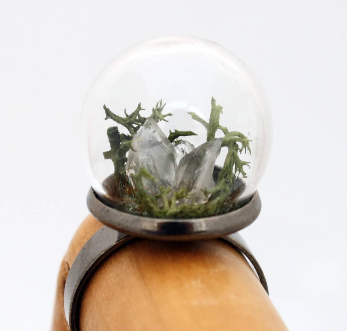 A wooden jointed hand displays this glass globe ring on a gunmetal ring base containing a crystal of lemurian seed quartz. The quartz is accompanied by reindeer moss and greenery inside the dome and displayed against a white background