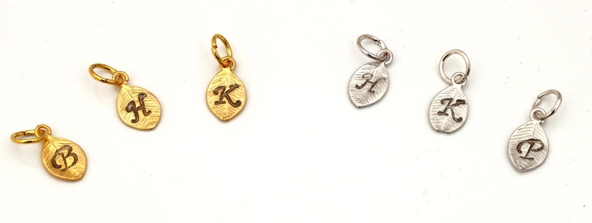 6 metal leaf-shaped charms with a small loop and jump ring on a white background. 3 of the charms are gold coloured with stamped calligraphic initials B, H and K. 3 of the charms are silver coloured with the stamped calligraphic initials H, K and P