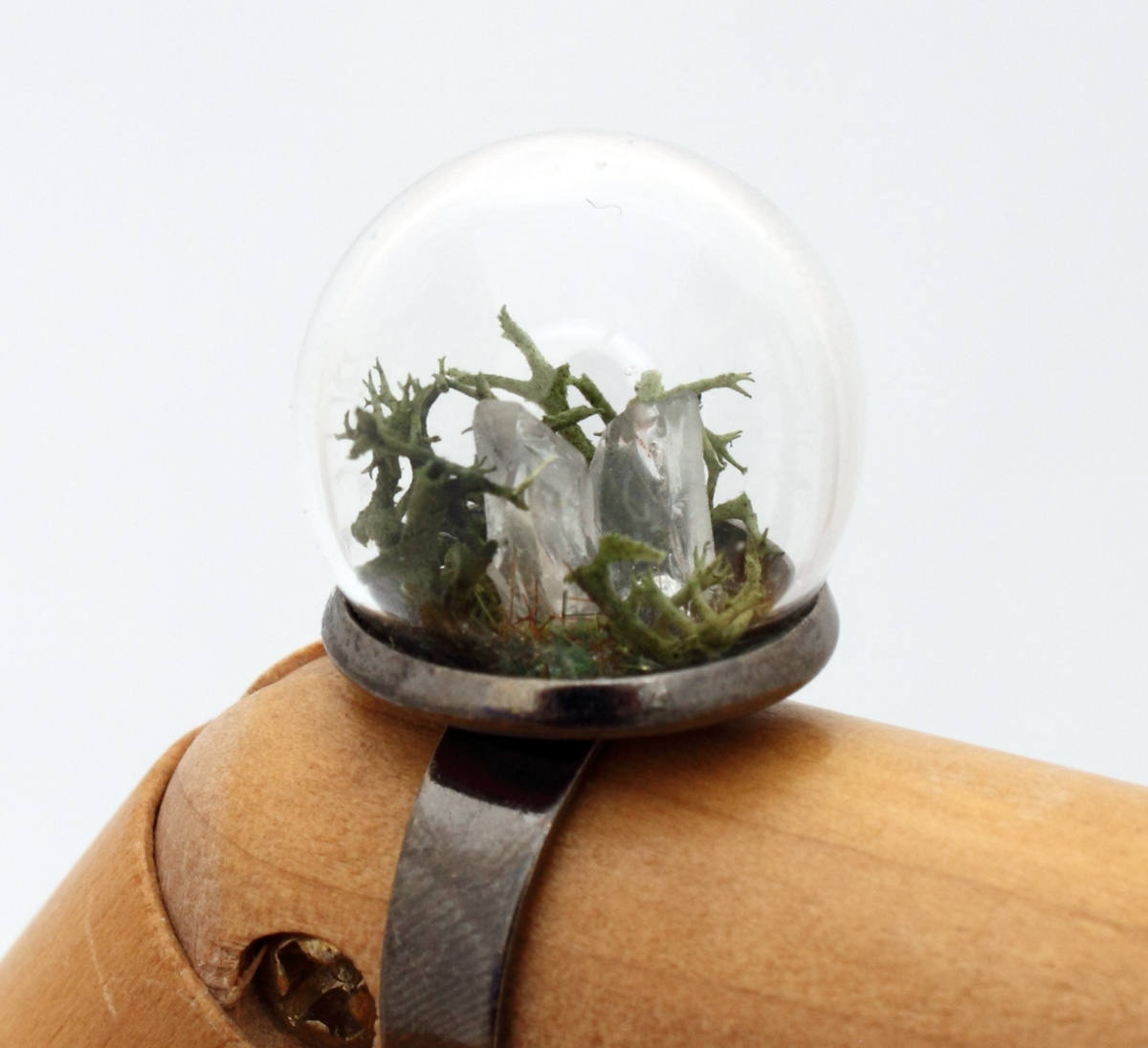 A wooden jointed hand displays this glass globe ring on a gunmetal ring base containing a crystal of lemurian seed quartz. The quartz is accompanied by reindeer moss and greenery inside the dome and displayed against a white background